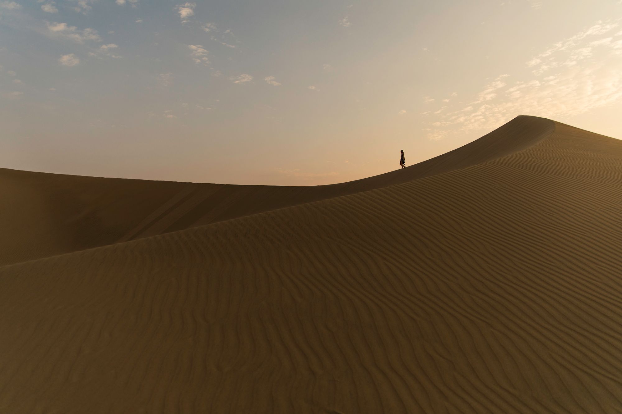 A lone figure walks along the crest of a large sand dune at dusk